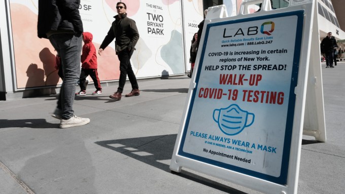 A Lab-Q sign advertising free walk-up COVID-19 testing sits on a public sidewalk. People walk in the background.