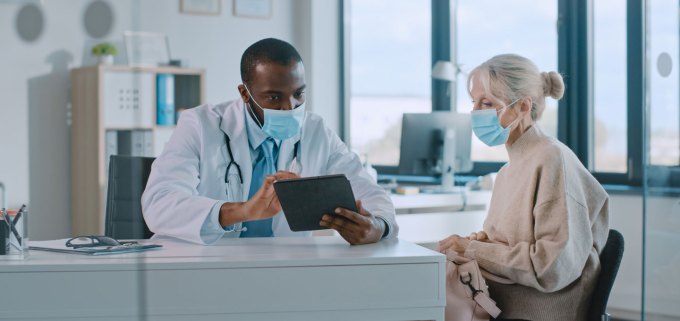 A patient sits next to a doctor both looking at something on a tablet