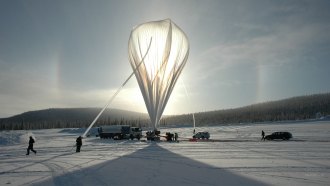 An upright weather balloon sits on a snowy landscape, surrounded by people and vehicles