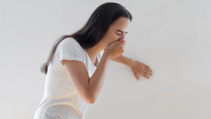 A woman with long dark hair wearing a white T-shirt holds her right hand over her mouth while her left hand braces her against the wall. The image conveys nauseousness.